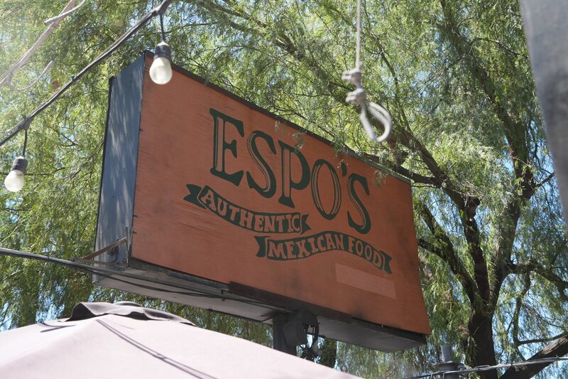 Hand-painted wooden Espo's sign hanging under a mesquite tree with string lights